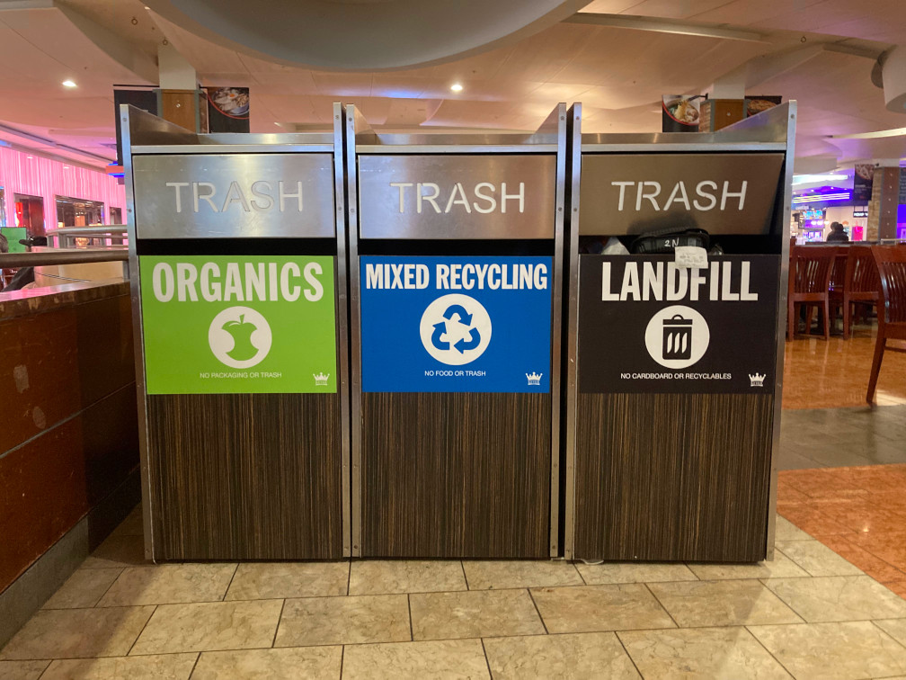 A unit of three rectangular waste receptacles in a shopping mall. The one on the left is labelled "organics" with white text on a green background, under the opening. Similarly, the one in the middle is labelled "mixed recycling" on a blue background, and the one on the right is labelled "landfill" on a black background. Despite the distinctions, the hinged doors where waste is deposited are all labelled "trash".